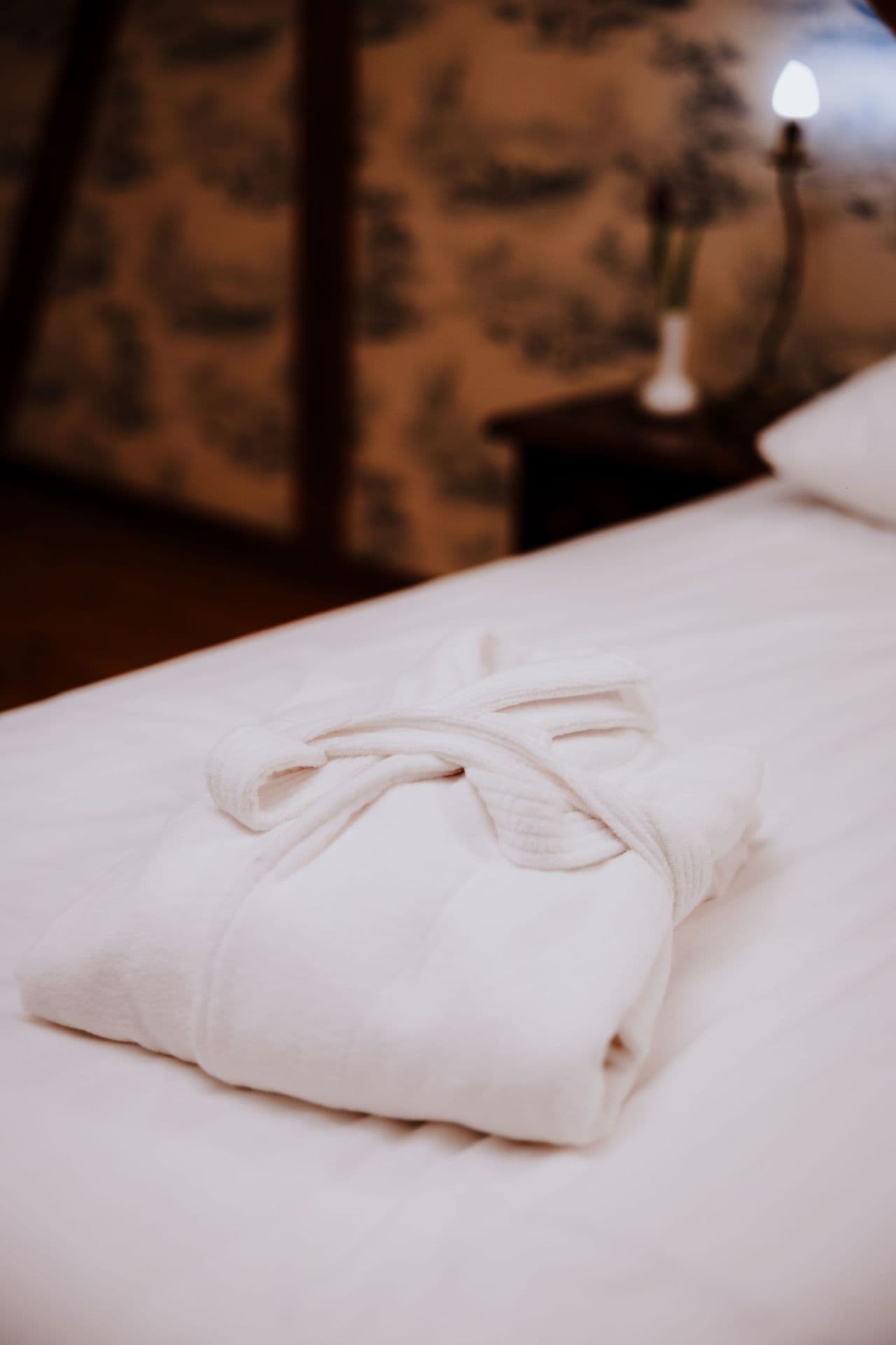 White folded bathrobe laid on crisp white bed linens, warm bedside lamp and patterned wallpaper.